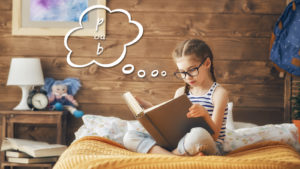 Uma menina jovem com óculos e tranças sentada na cama, concentrada na leitura de um livro. Ela está em um quarto aconchegante com paredes de madeira e decorações infantis, como uma luminária e um boneco. O cenário é tranquilo e ideal para estudos ou leitura em casa.